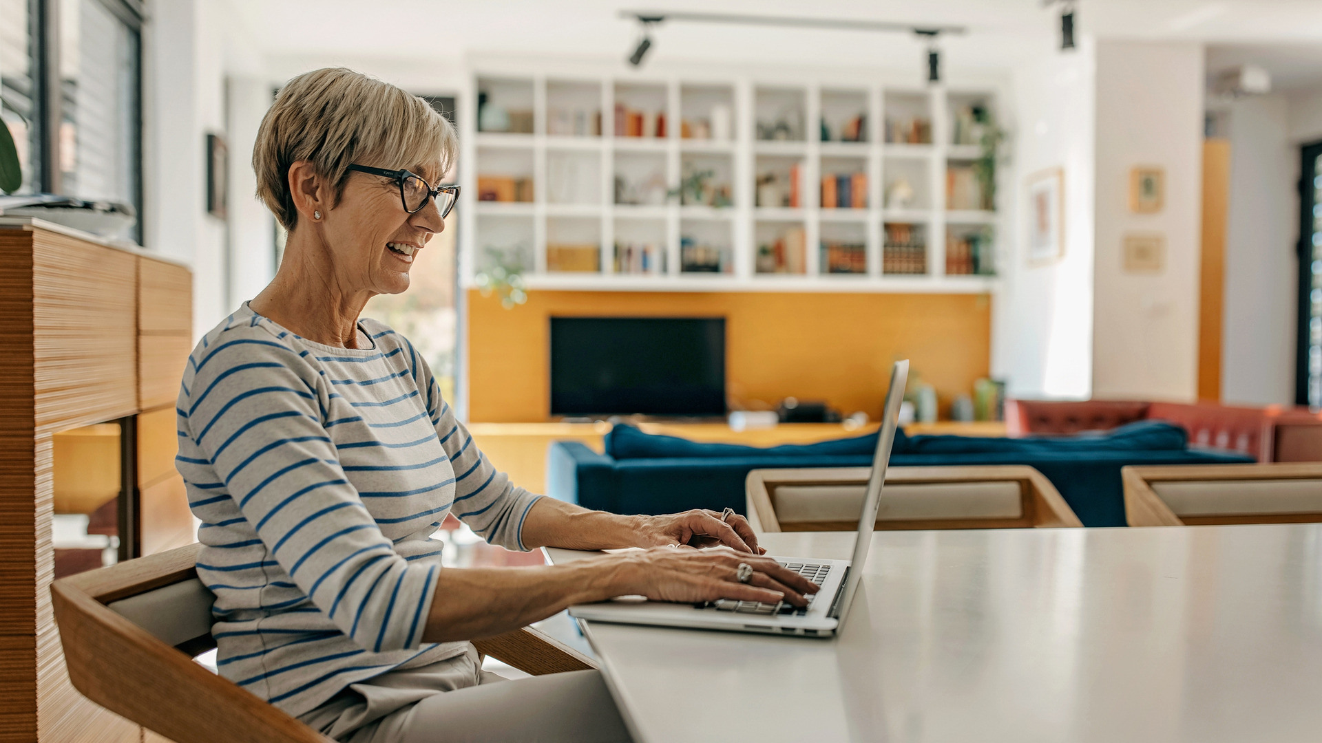 Middelaldrende kvinne med kort grått hår sitter ved kjøkkenbordet og ser på PC-skjermen