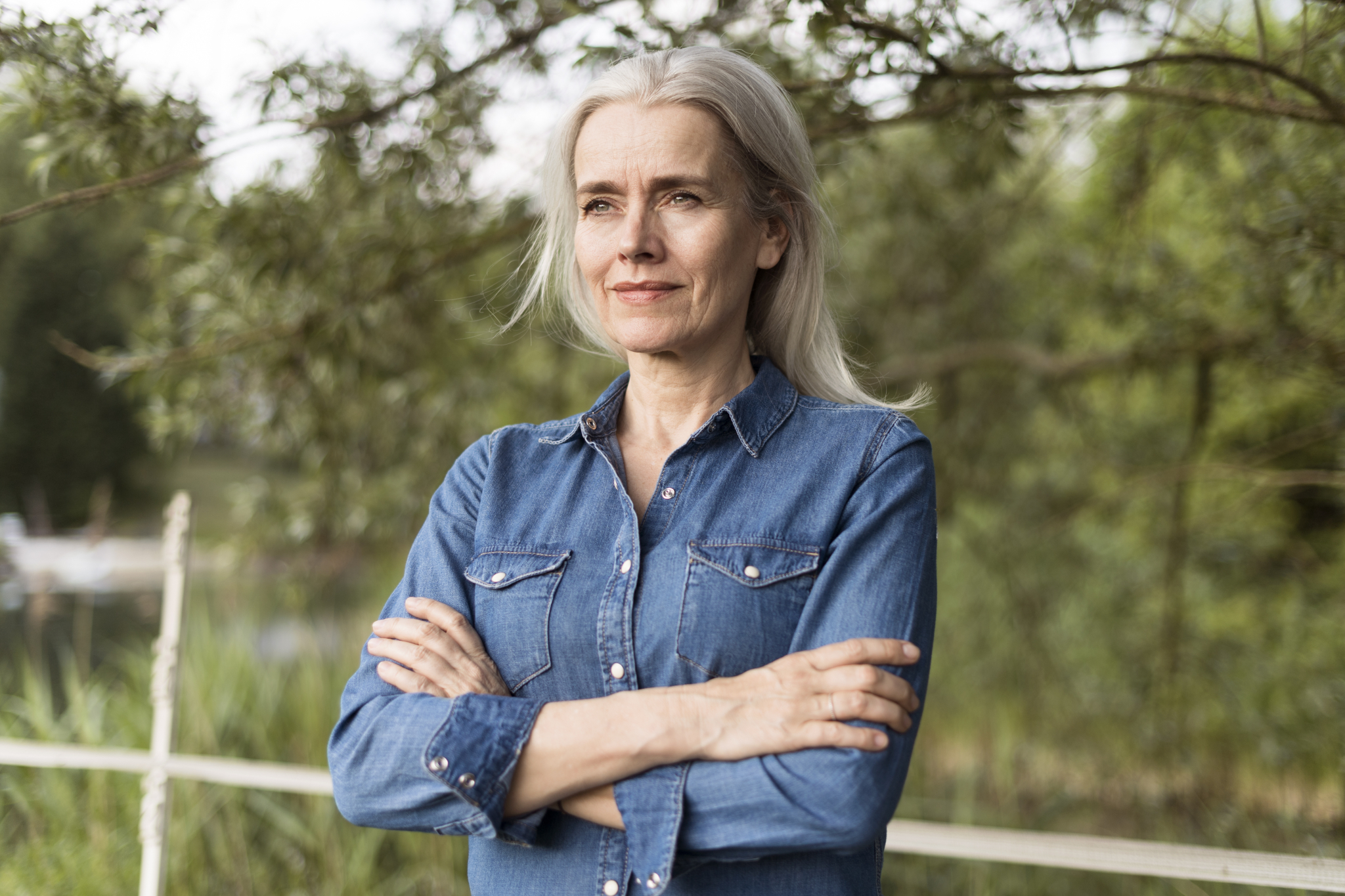 Moden kvinne i olaskjorte og grått hår står ute med hendene i kors over brystet, ser bestemt ut.