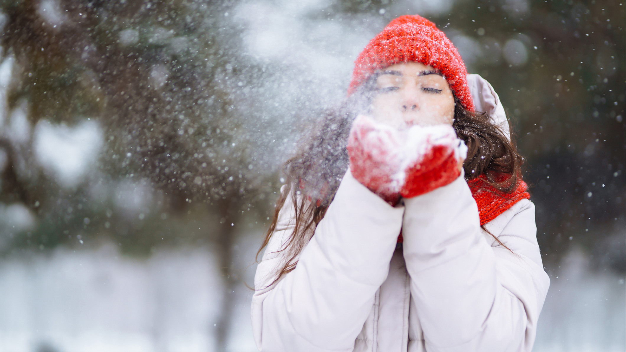 Kvinne i rød lue og røde vanter blåser snø mot kamera