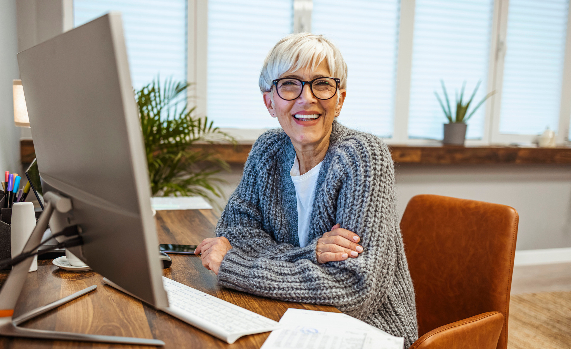 Kvinne med grått hår og briller smiler bredt mot kamera der hun sitter ved pulten sin.