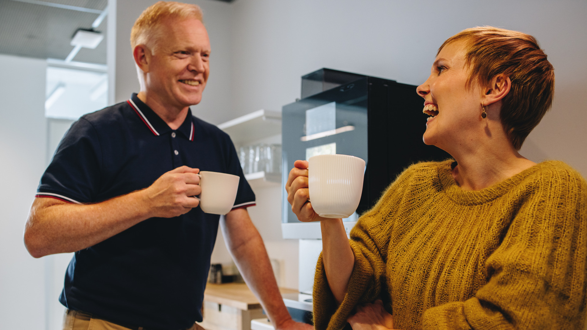 En mannlig og kvinnelig kollega konverserer foran kaffeautomaten på jobben